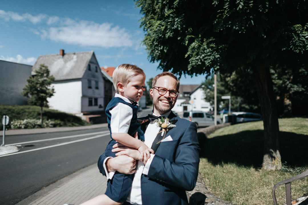 Hochzeit-im-Heyligenstaedt Bräutigam mit kleinem Jungen auf dem Arm vor der Kirche Kinzenbach
